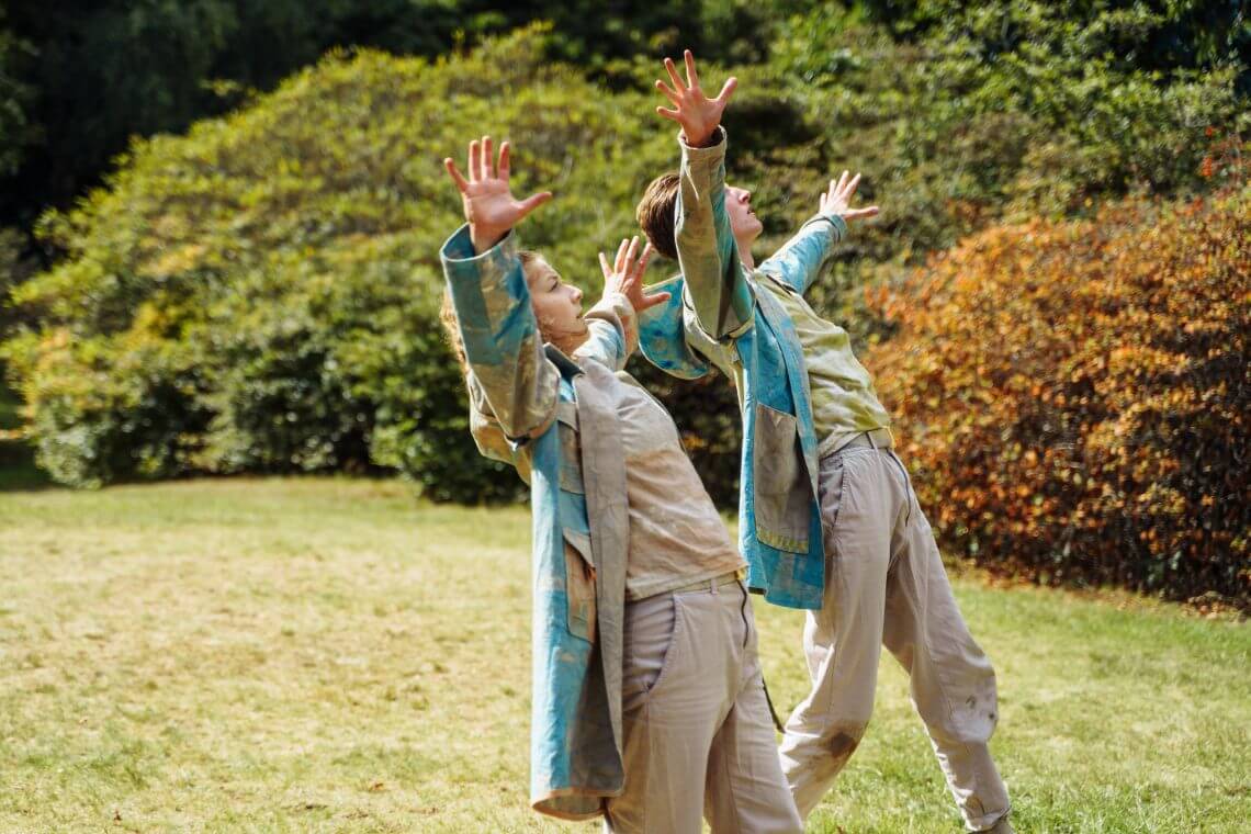 Two dancers as if blown backward by a gust of air open both arms up to the sky. Outside on grass.