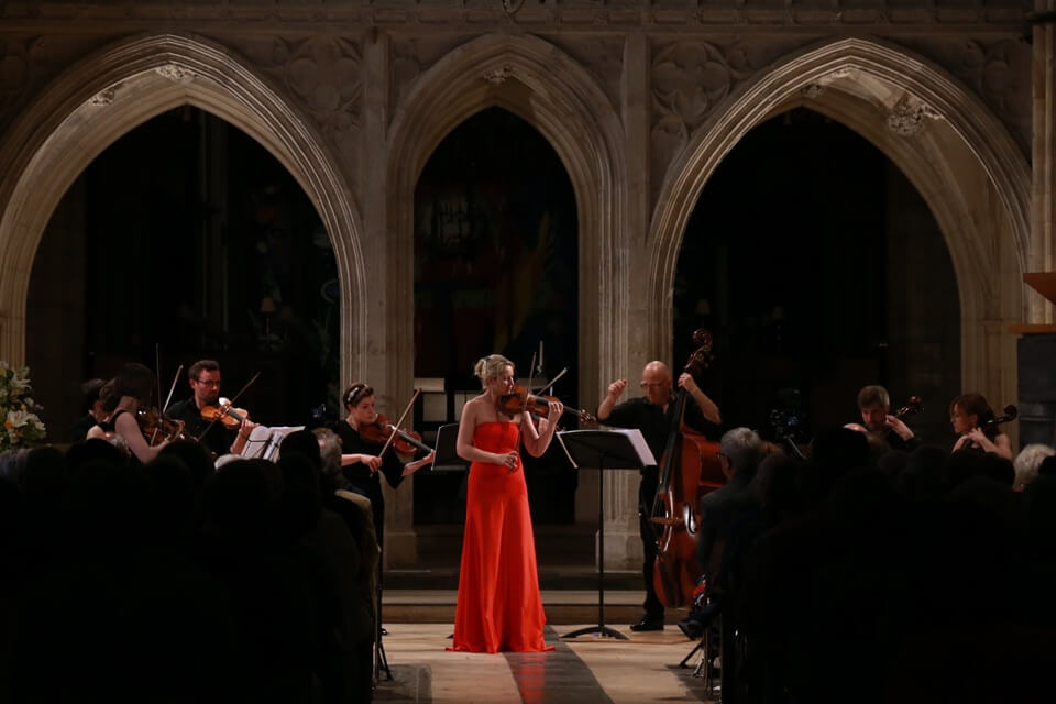female violinist in red gown surrounded by audience and other performers in a chapel