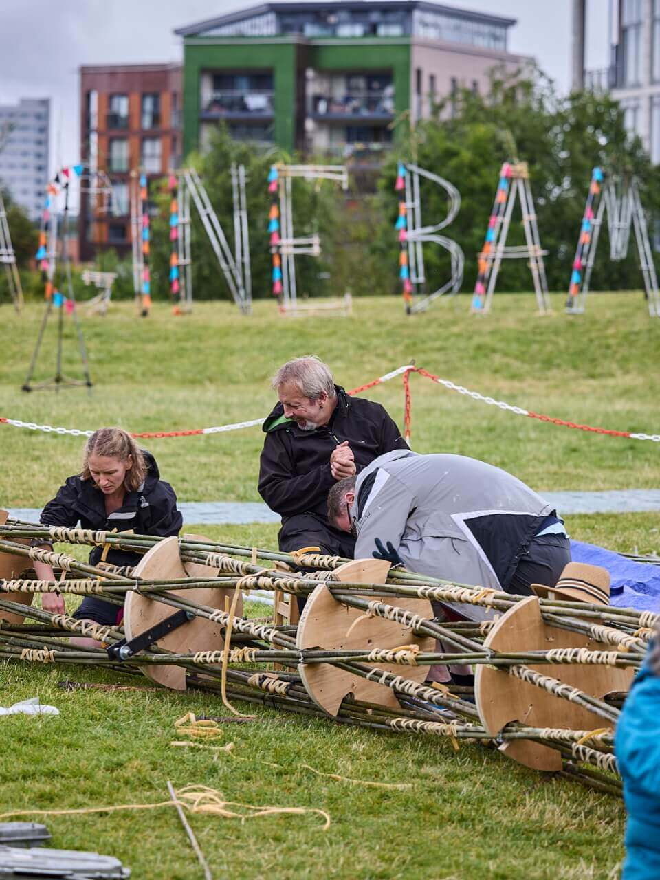 Image features 3 people sitting on the grass tying bamboo together to create the arch structure. In the background you can see some large bamboo letters, they spell out Imagine Bamboo however you can only see part of the wording on this image, you can read GINE and BAM.