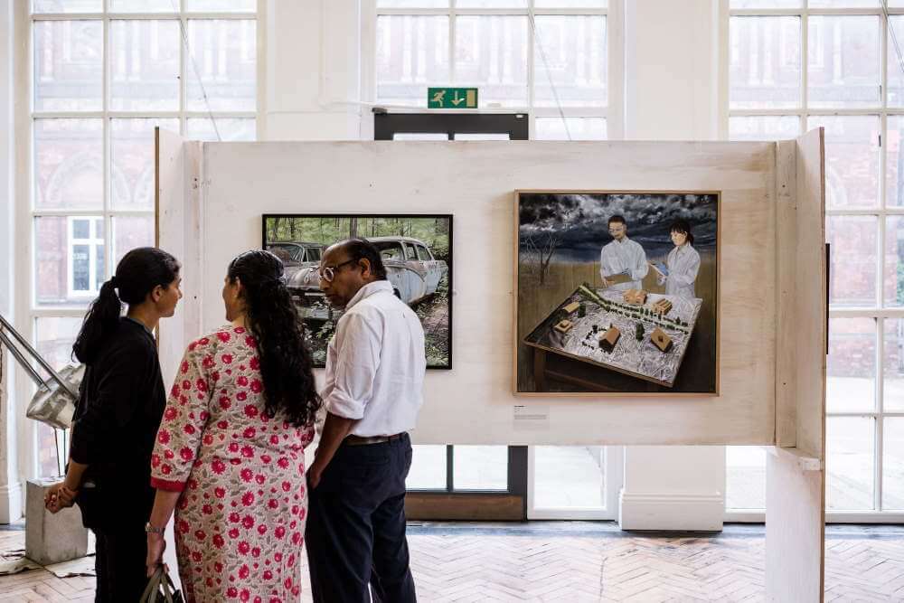Group of guests standing and talking to the left side of the image with a board in the background featuring two of the works in the exhibition.