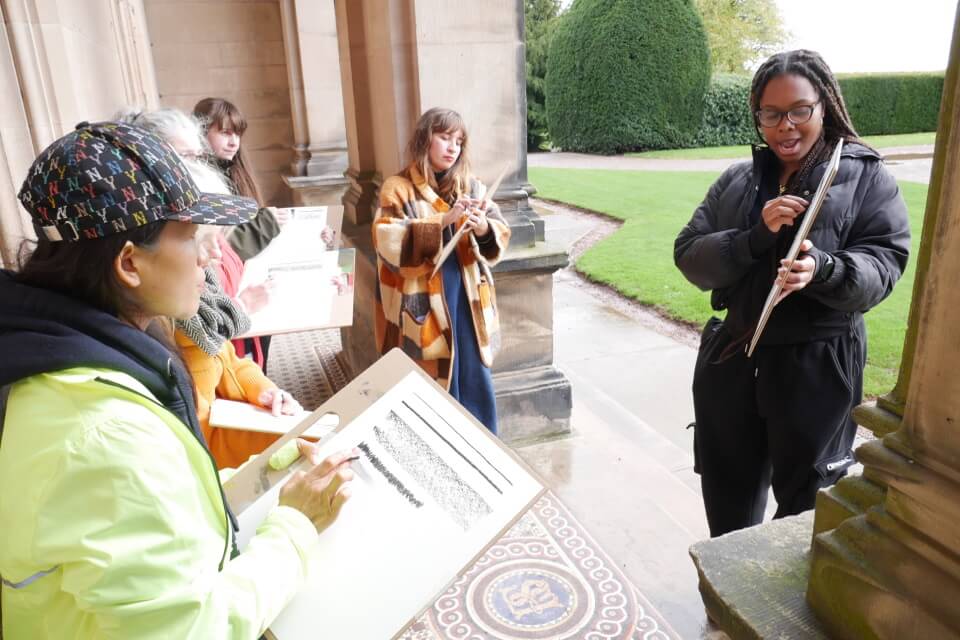 Group of people holding drawing boards with paper, participating in a charcoal drawing workshop taking place outdoors