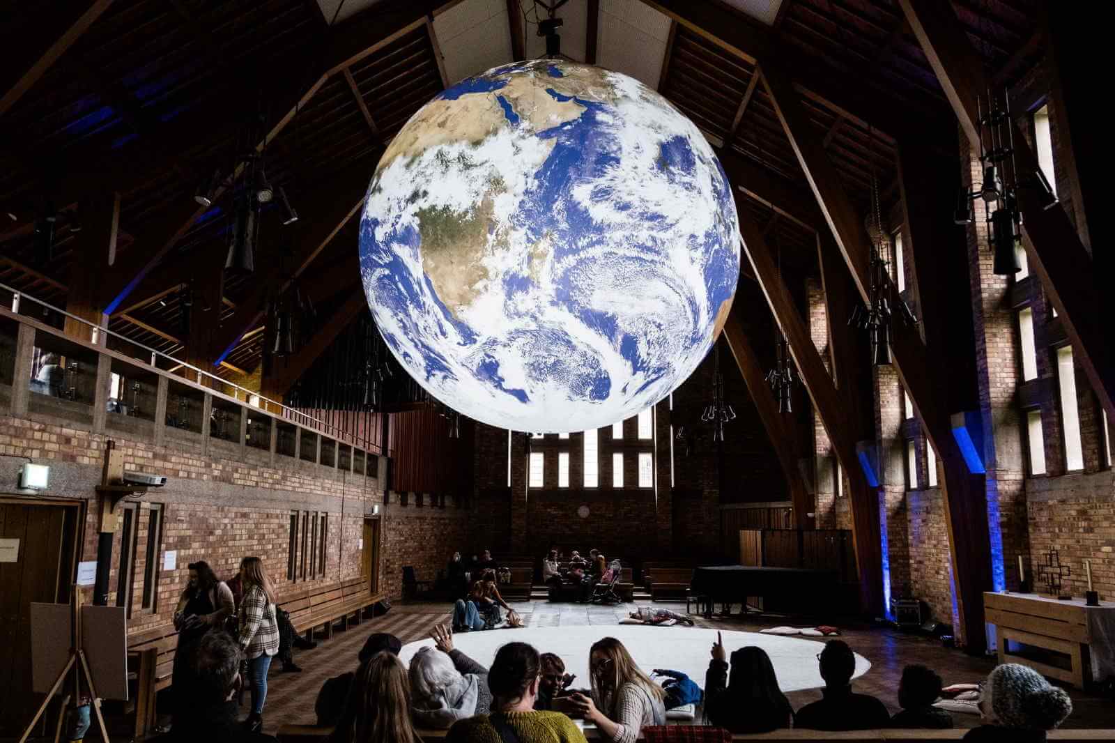 image credit Jenny Harper. Main image large earth over crowd of people inside Chapel space. Second image row of people watching an aerial artist swing on an arch made of bamboo outdoors with trees in the background. 