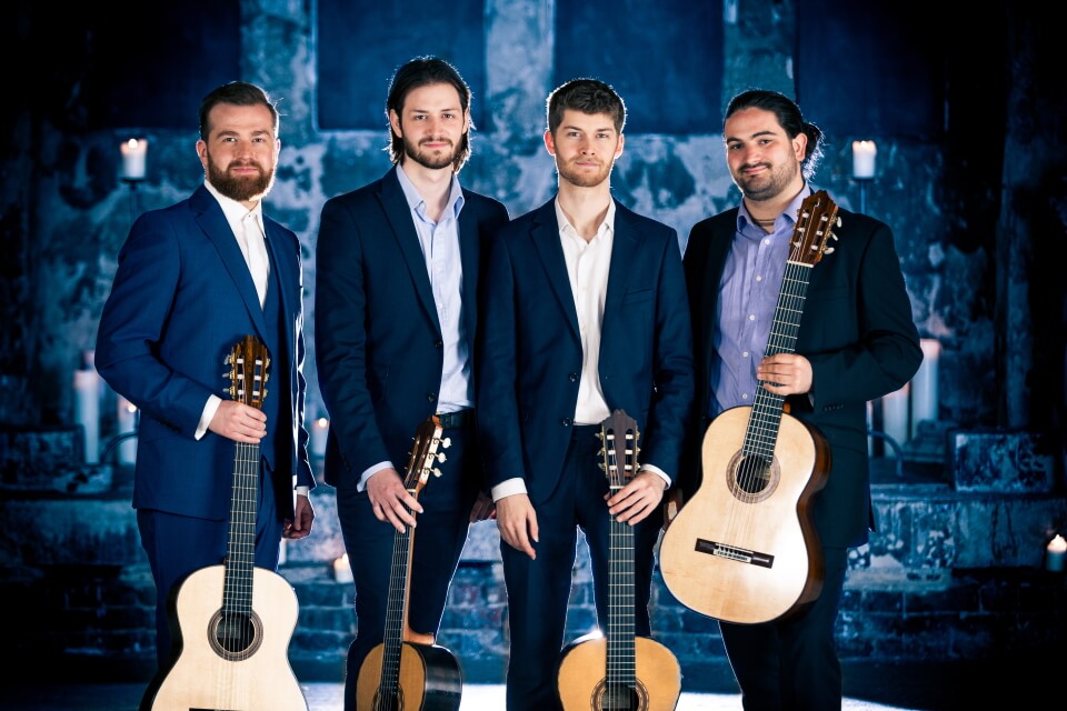 image of guitar quartet holding their guitars inside a blue toned church with candles