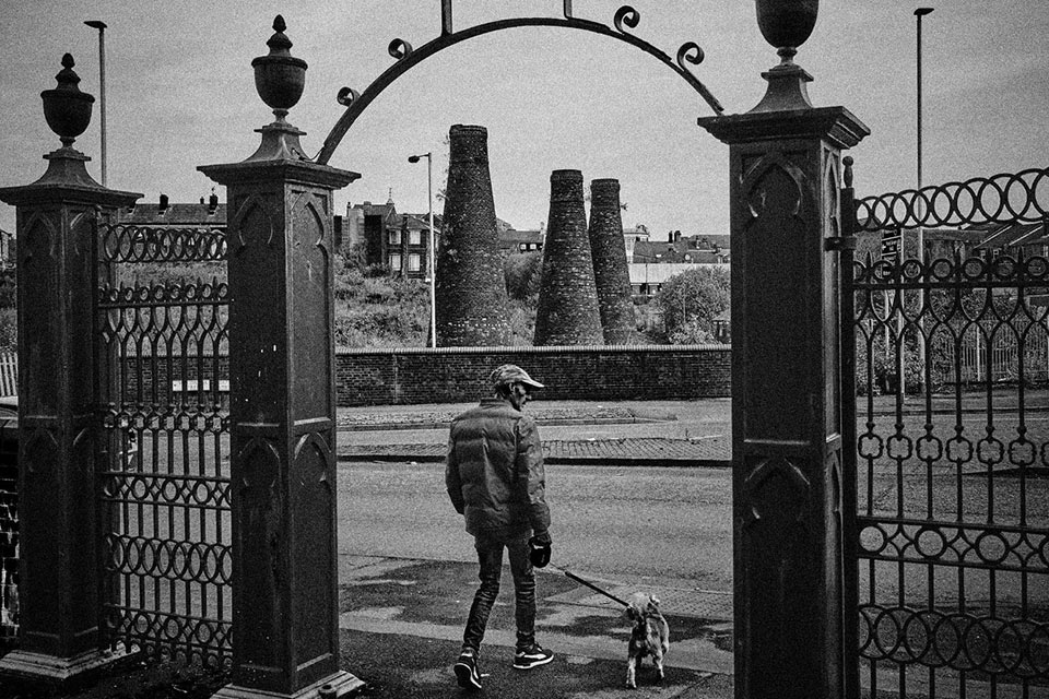 black and white photograph of a close up of a bottle kiln, a man with a dog walk under an iron arch