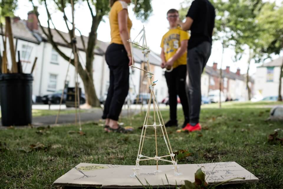 Image of people standing with mini bamboo structure in foreground placed on top of paper on grass