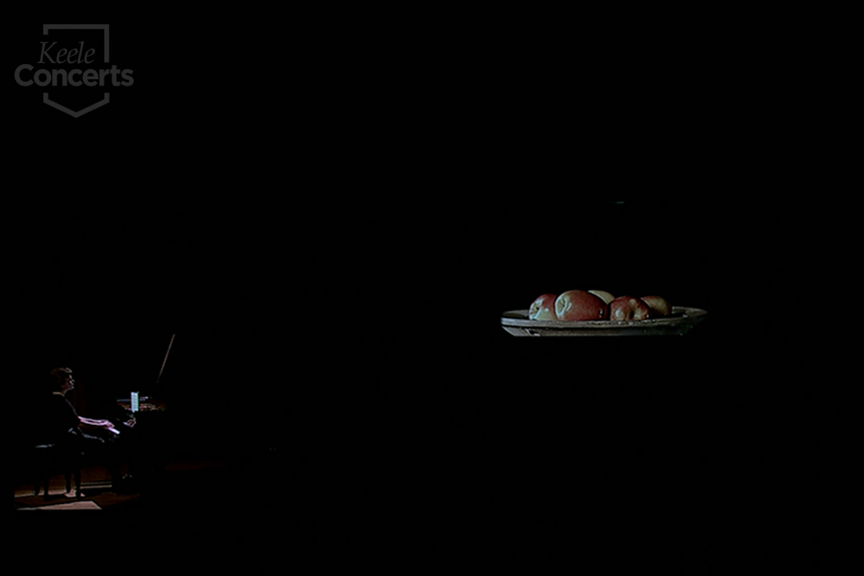 Female performer sat at piano in dark space with projection of apples on a plate.