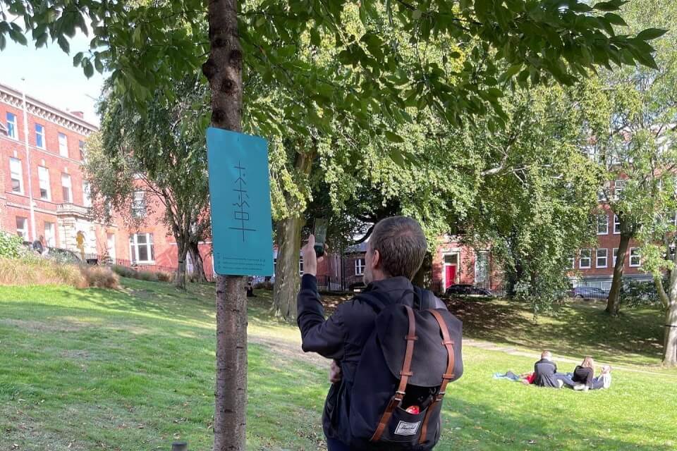 a person holding a phone towards a tree with a large blue plaque with icons on it, in a park. 