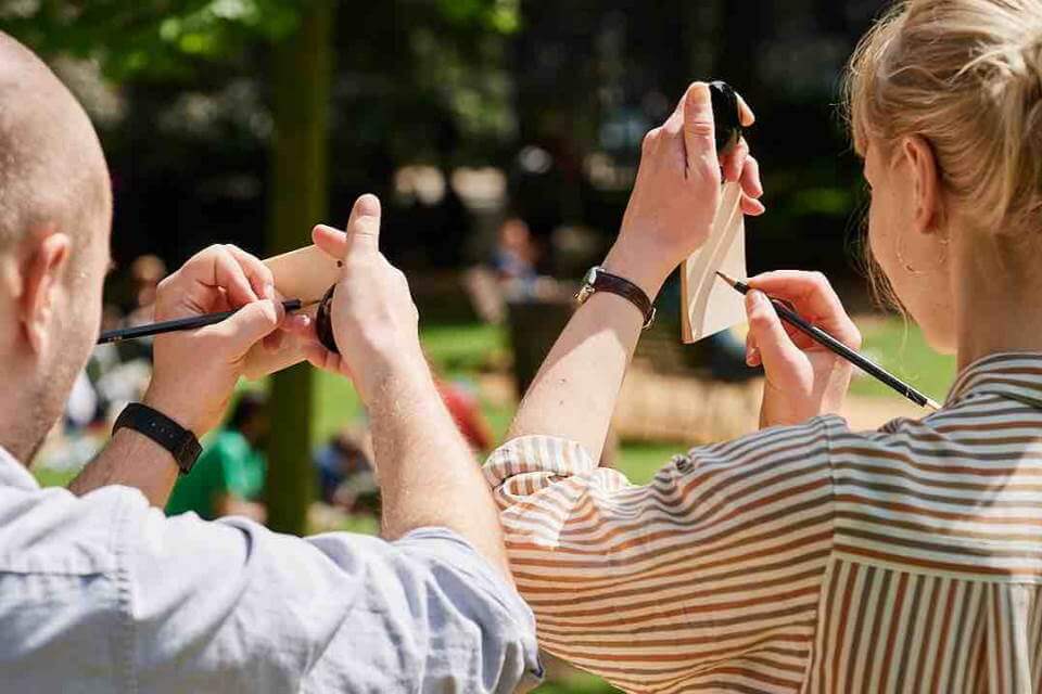 two people holding a small mirror and drawing on a small pad at the same time outside