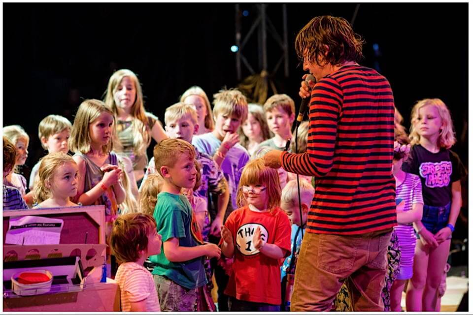 a group of children dancing surrounding a person wearing a red and black striped top holding a mic