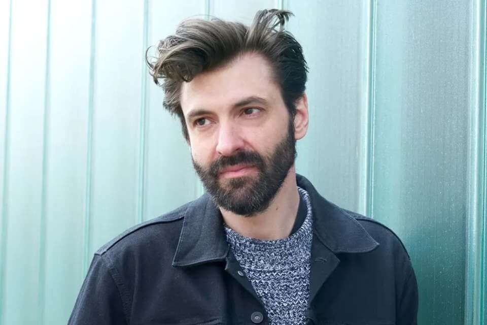 image of man wearing charcoal jacket and blue fleck jumper, brown hair swept to one side, and beard.