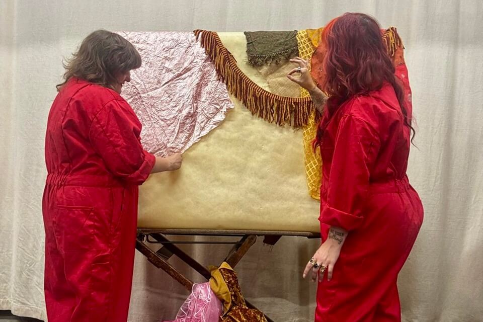 two people dressed in red boiler suits with different fabrics on a board