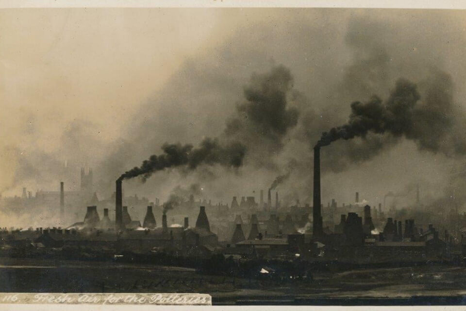 black and white photo of potteries landscape, multiple chimneys puffing out smoke and bottle kilns.