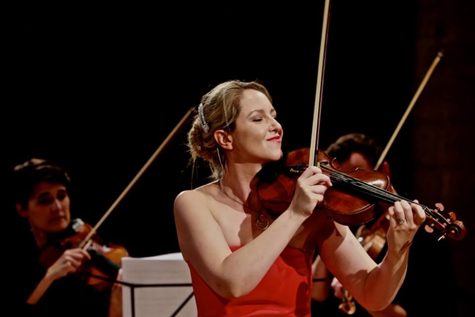 A lady in a red dress holding a violin, plucking the strings and smiling