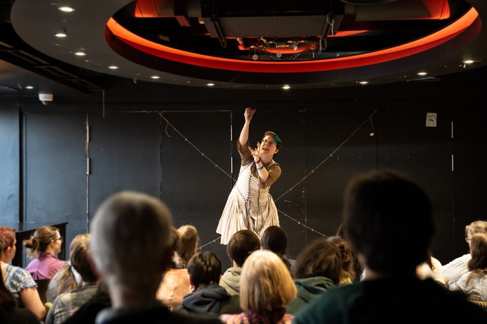 a performer centre stage reaching to the sky, audience members in the foreground