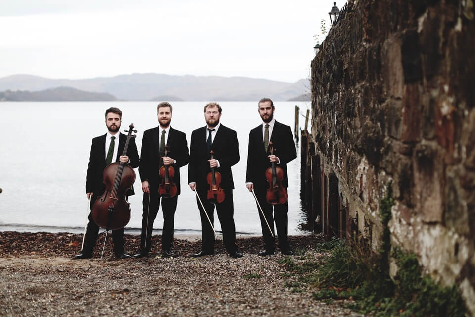 A photographic portrait of 4 musicians and their string instruments in front of a misty lake.