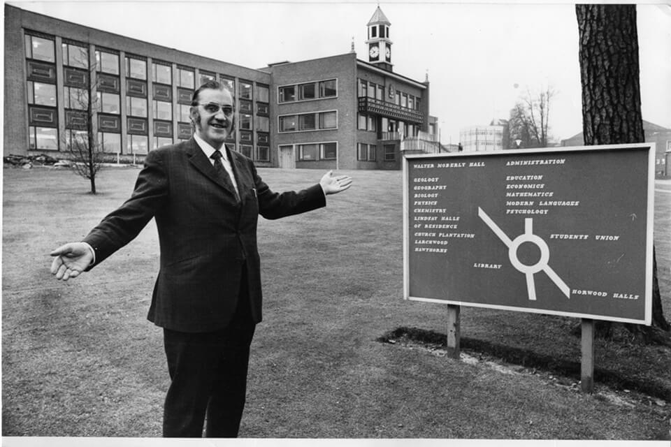 man standing infront of university building gesturing towards a campus roadsign