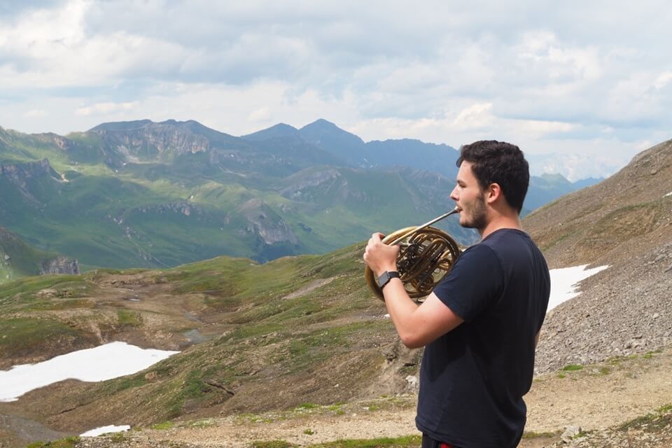 male horn player standing on a mountain playing across a valley