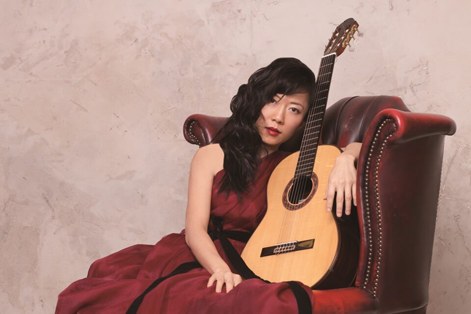 Xuefei seated wearing deep red gown, leaning against her guitar