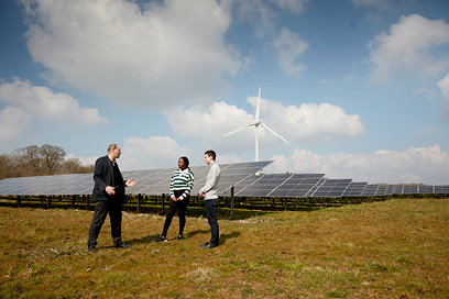  Two students and a lecturer giving a talk with solar panels and wind turbines in the background 