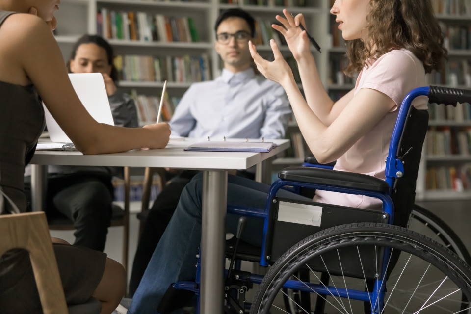 People sat discussing around a table in a library-setting. One of the participants is in a wheelchair.