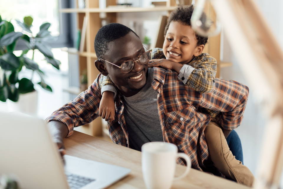 An adult and a child laughing together, while the adult works on a laptop computer.