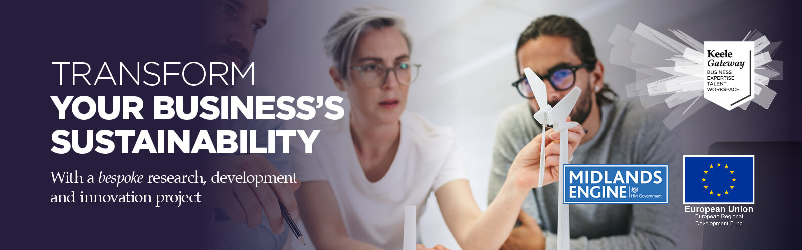 Photograph of three people standing at a workbench in discussion, pointing to models of a wind turbine