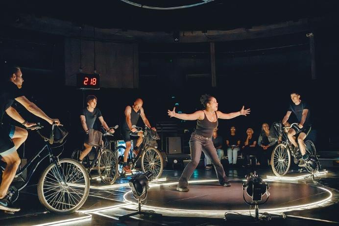 A woman stands on a darkened theatre stage with cyclists on bicycles surrounding her.