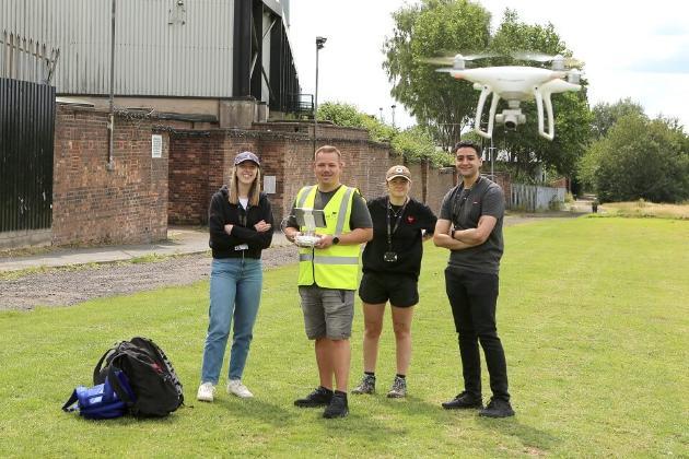 Four people standing on a green space controlling a drone with a remote.