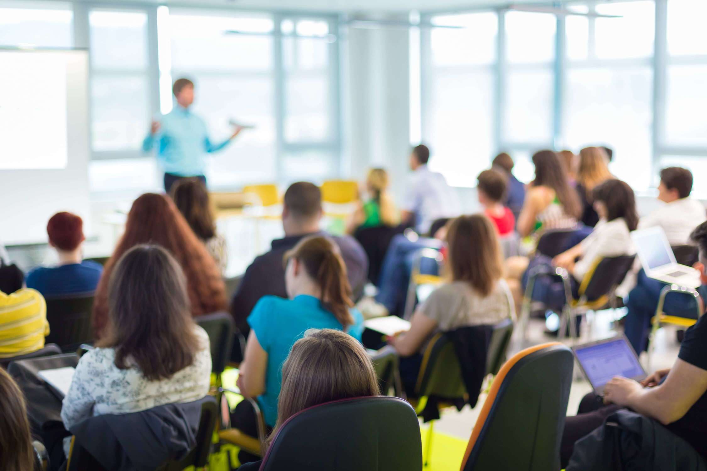 Room of students sat at desks, with backs to the camera, and a male teacher presenting at the front 