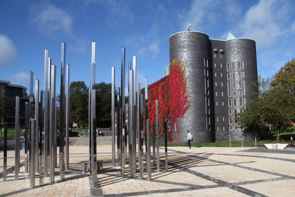 The exterior of the Chapel viewed from Union Square, with the metal Forest of Light Sculpture in the foreground.  The chapel is a striking blue brick building with two rounded towers at the near end.  The left hand side of the building is covered in Virginia creeper which has leaves ranging from green to vivid red.