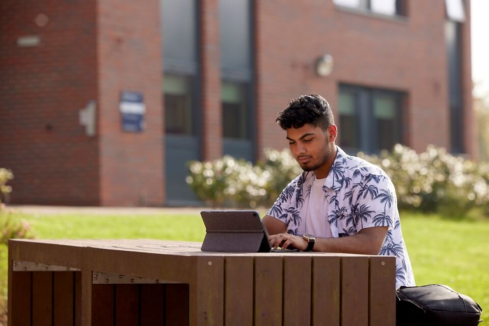 Student sitting outside Barnes accommodation on Keele campus, looking at laptop