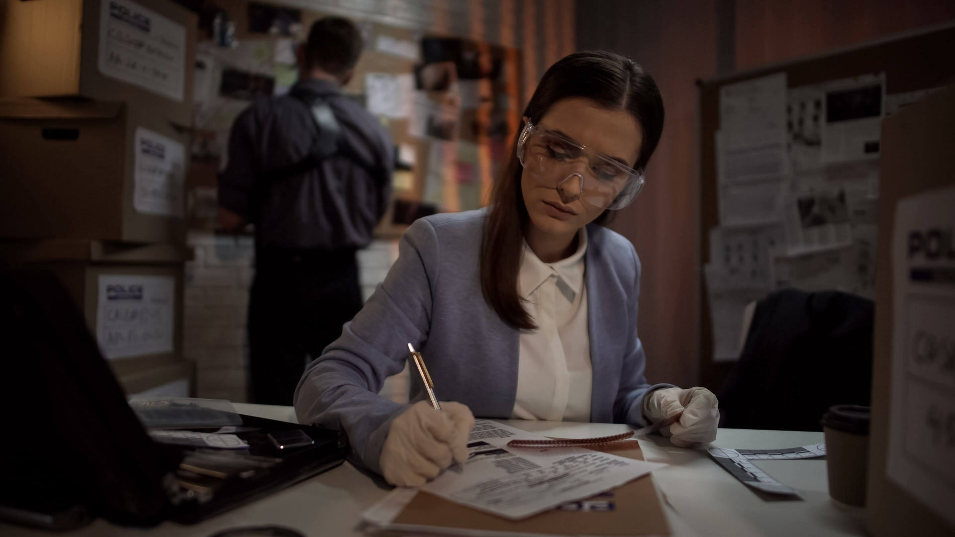 A female investigator in protective glasses sat at a table writing a report with a male in the background looking at evidence boxes