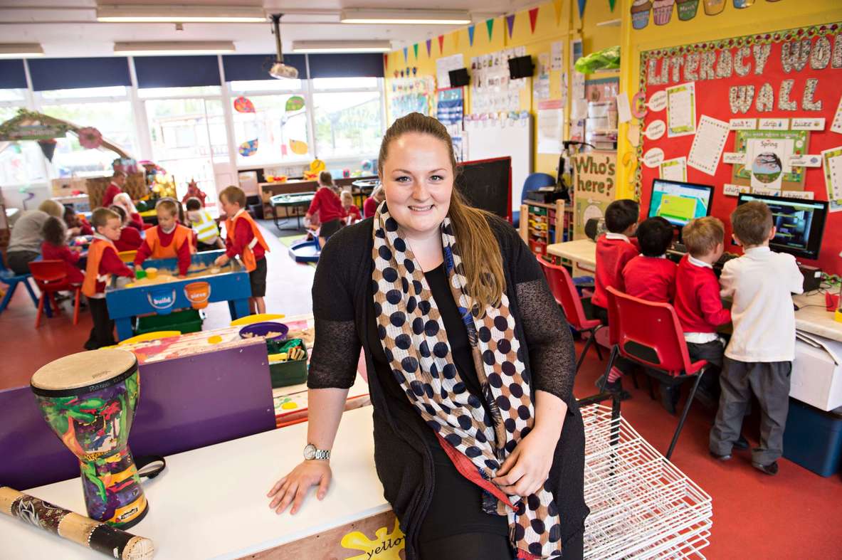 Female teacher sat smiling at the front of a classroom with young primary school children working in the classroom 