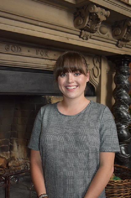 Female in grey dress standing front of big fireplace smiling at the camera 