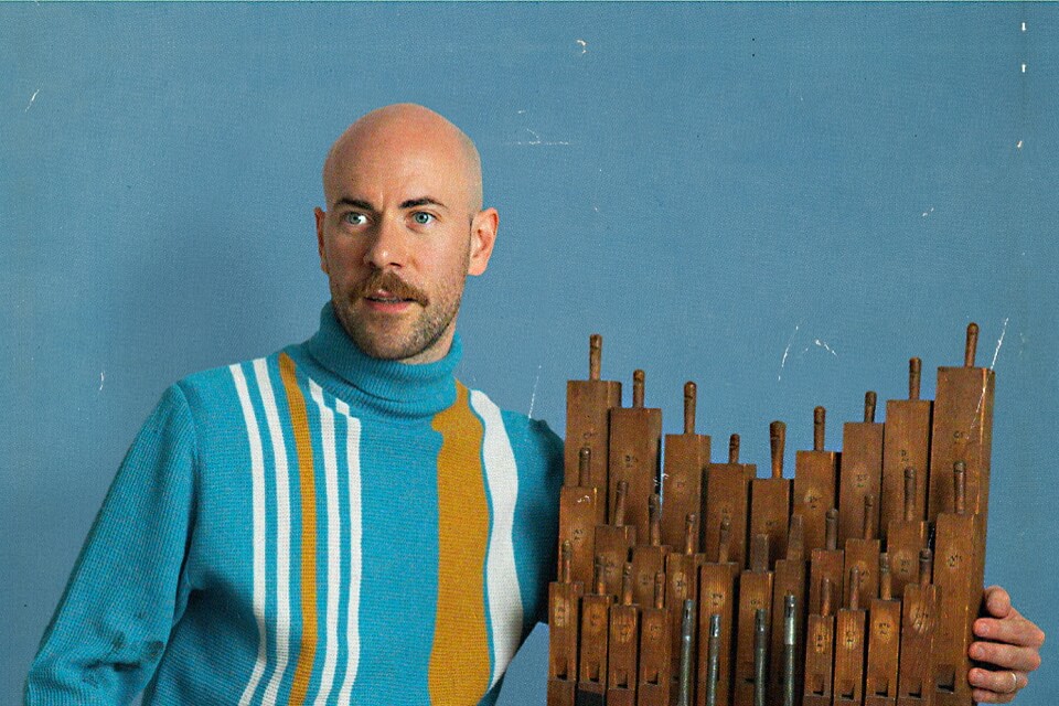 image of Brad Field wearing turquoise jumper with orange and white stripes and arm around an organ