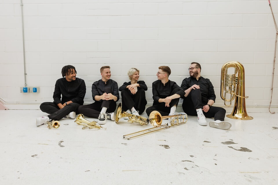 five people wearing black sat in a white space with brass insruments on the floor happily chatting