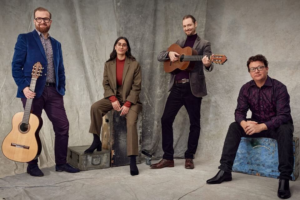 Photograph of four musicians sitting on boxes, two with guitars, all looking to camera.